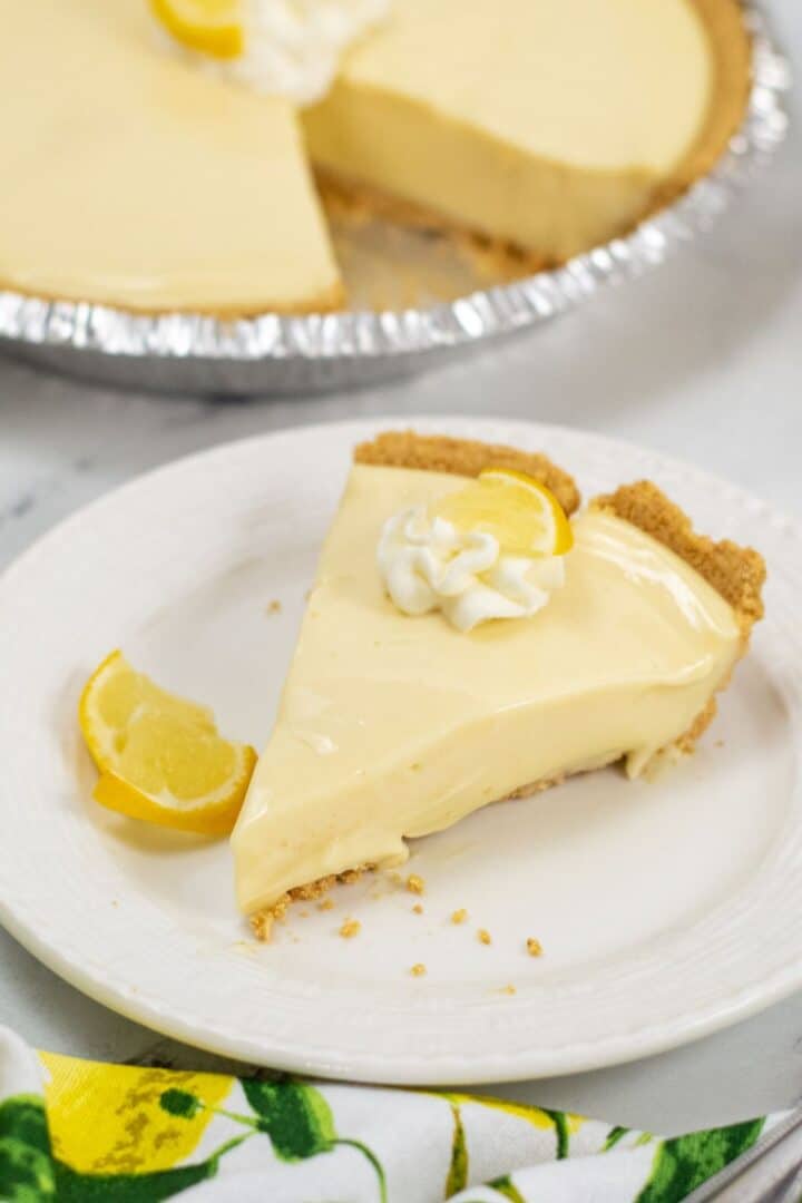 A slice of creamy Lemon Icebox Pie with a graham cracker crust, topped with whipped cream and a lemon wedge, sits on a white plate; the rest of the pie is visible in the background.