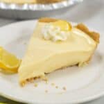 A slice of Lemon Icebox Pie with a graham cracker crust, topped with whipped cream and a lemon wedge, served on a white plate.