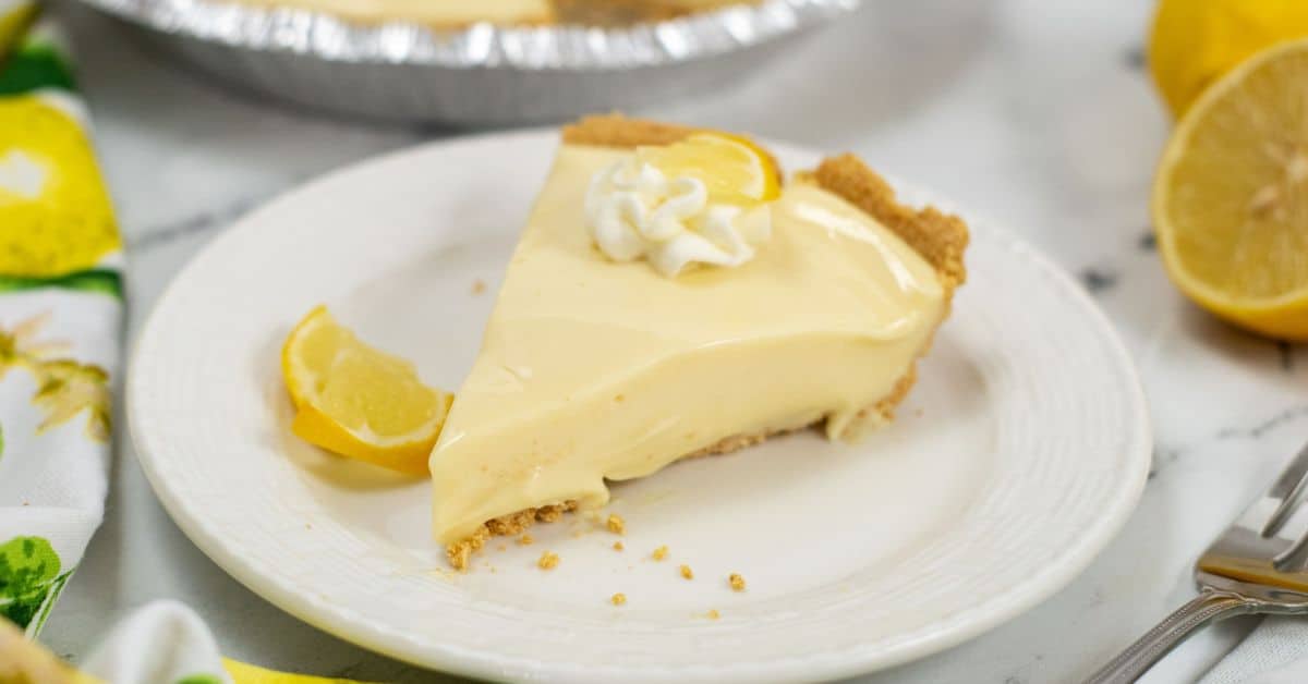 A slice of Lemon Icebox Pie with a graham cracker crust, topped with whipped cream and a lemon wedge, served on a white plate.
