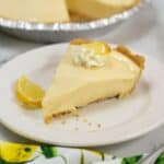 A slice of Lemon Icebox Pie with a graham cracker crust, topped with whipped cream and a lemon wedge, sits on a white plate. The whole pie is in the background.