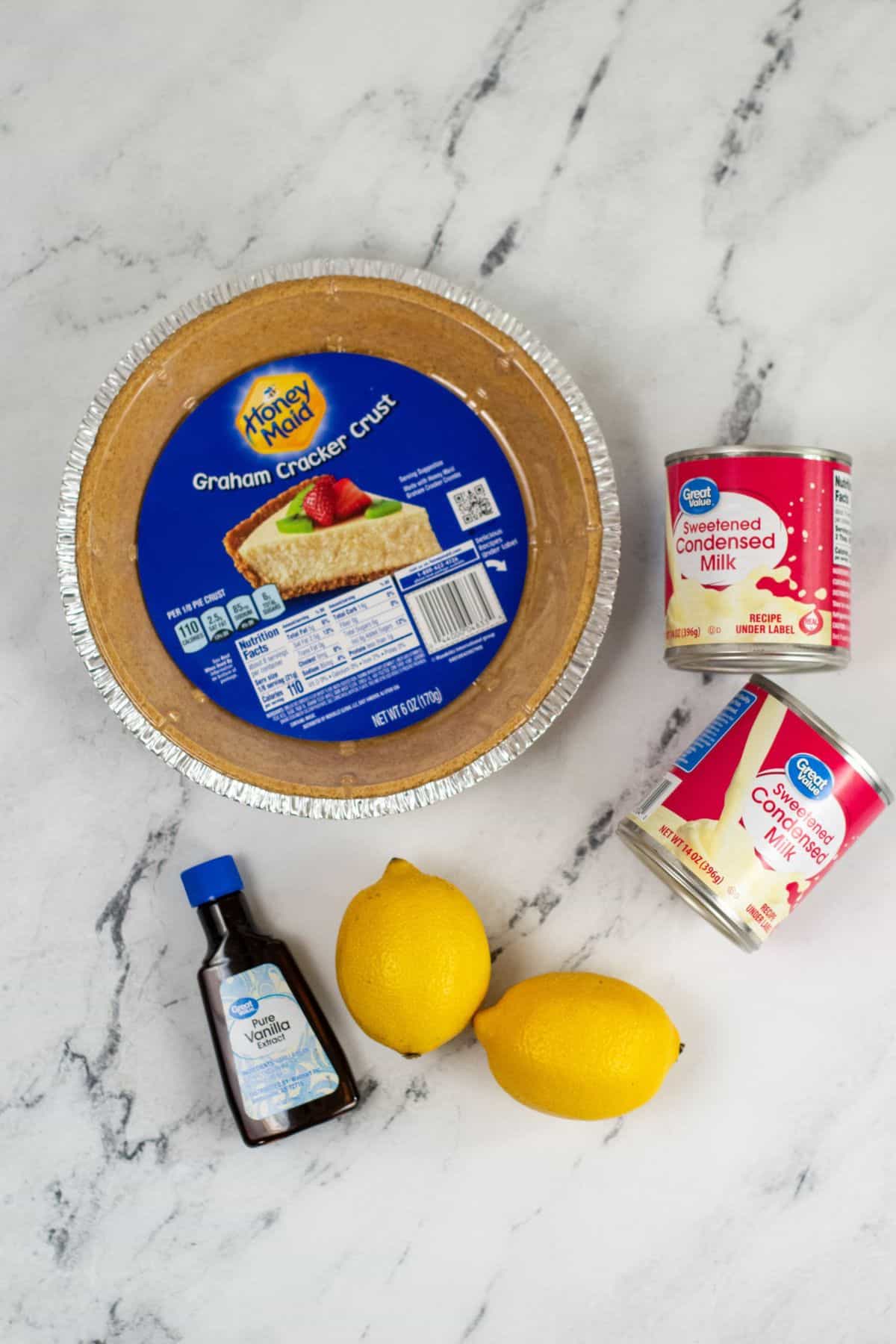 A graham cracker pie crust, two cans of sweetened condensed milk, a bottle of vanilla extract, and two lemons arranged on a white marble surface.