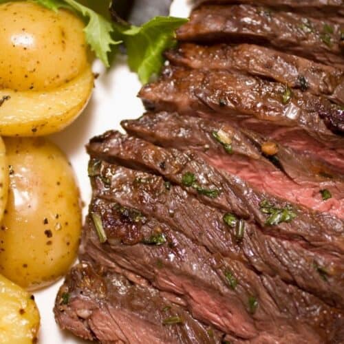 Marinated Skirt Steak, sliced medium-rare and garnished with herbs, served with roasted baby potatoes and mixed greens on a white plate.