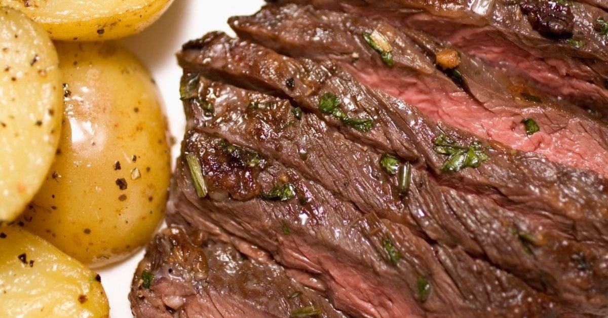 Marinated Skirt Steak, sliced and grilled with visible herbs, served alongside seasoned roasted potatoes on a white plate.