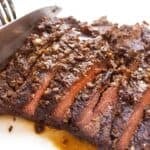 A knife is slicing a cooked, seasoned marinated skirt steak on a white cutting board, with visible juices glistening beneath the tender meat.