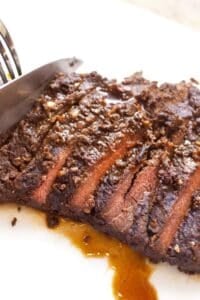A knife is slicing a cooked, seasoned marinated skirt steak on a white cutting board, with visible juices glistening beneath the tender meat.