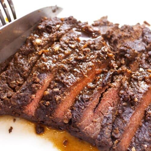 A knife is slicing a cooked, seasoned marinated skirt steak on a white cutting board, with visible juices glistening beneath the tender meat.