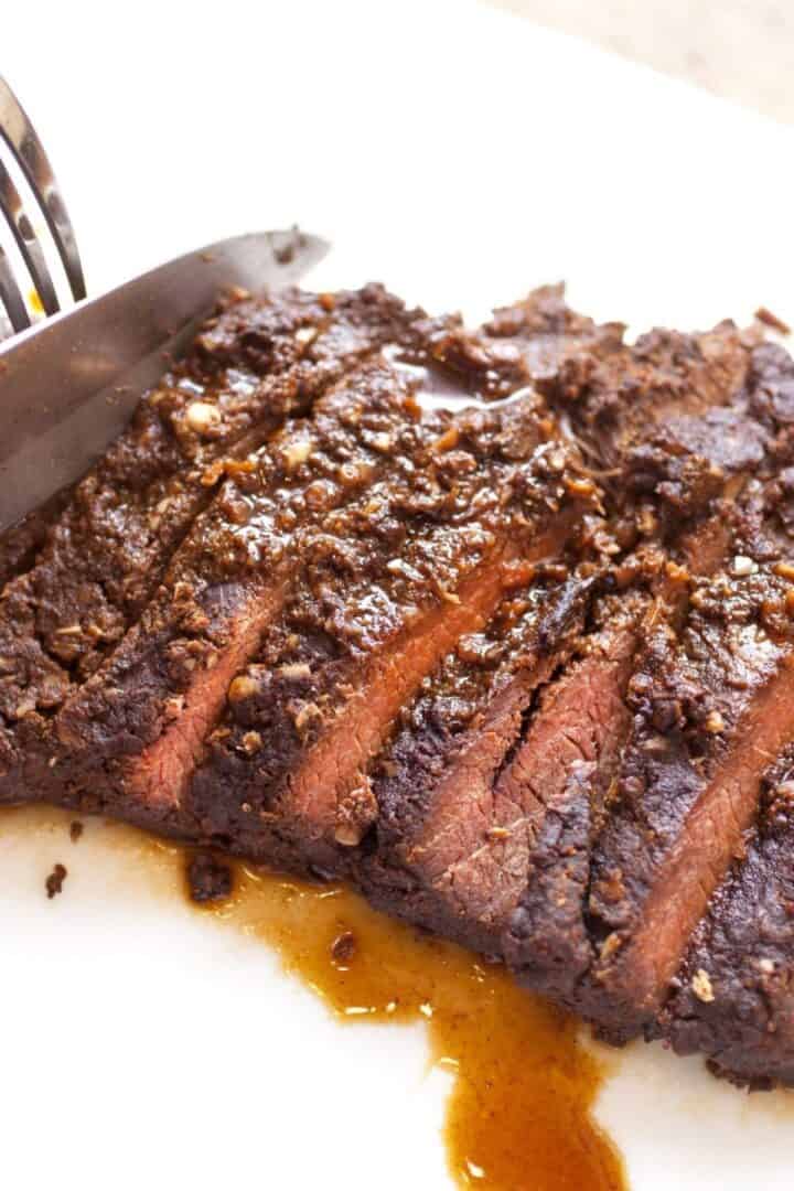 A knife is slicing a cooked, seasoned marinated skirt steak on a white cutting board, with visible juices glistening beneath the tender meat.