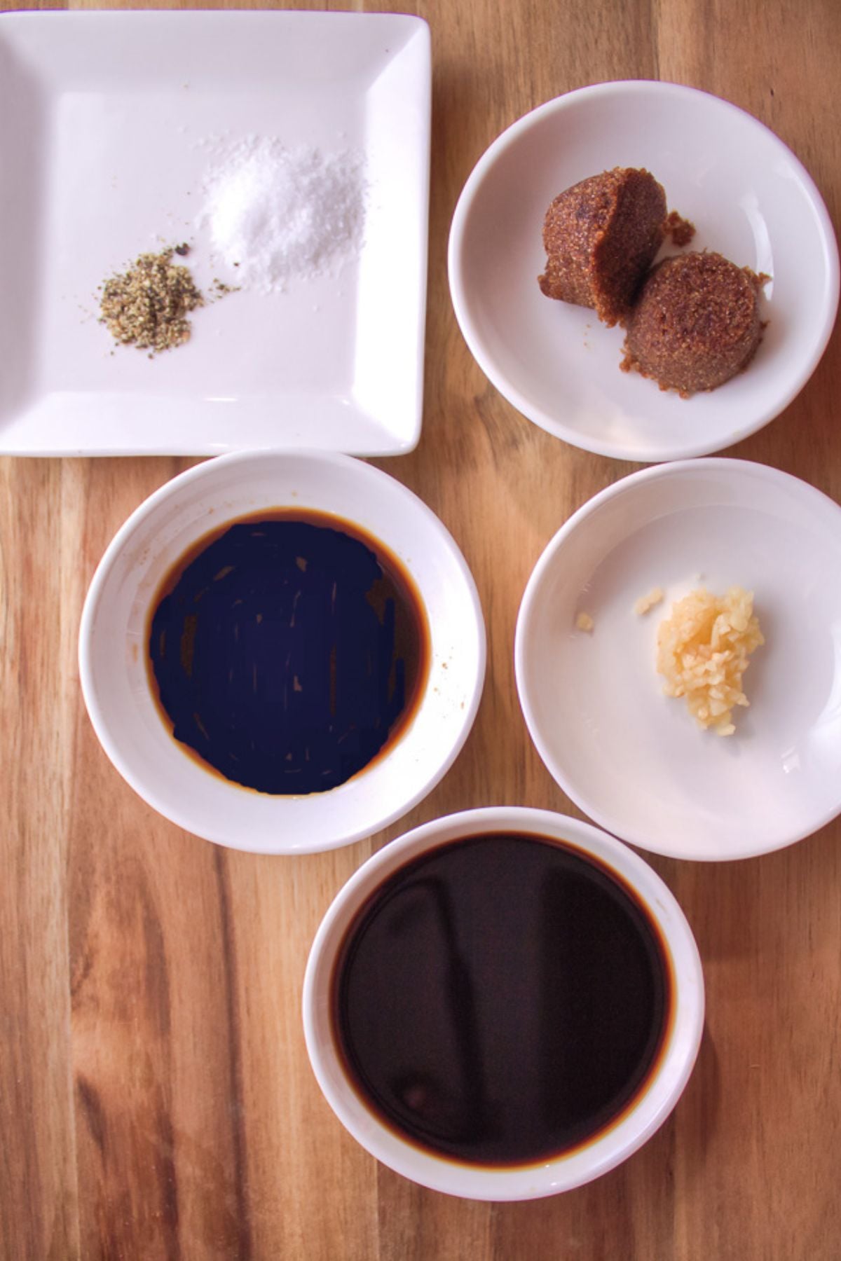 Five small bowls and plates,  hold ground pepper with salt, brown sugar, soy sauce, minced garlic, and another dark liquid—all beautifully arranged on a wooden surface.