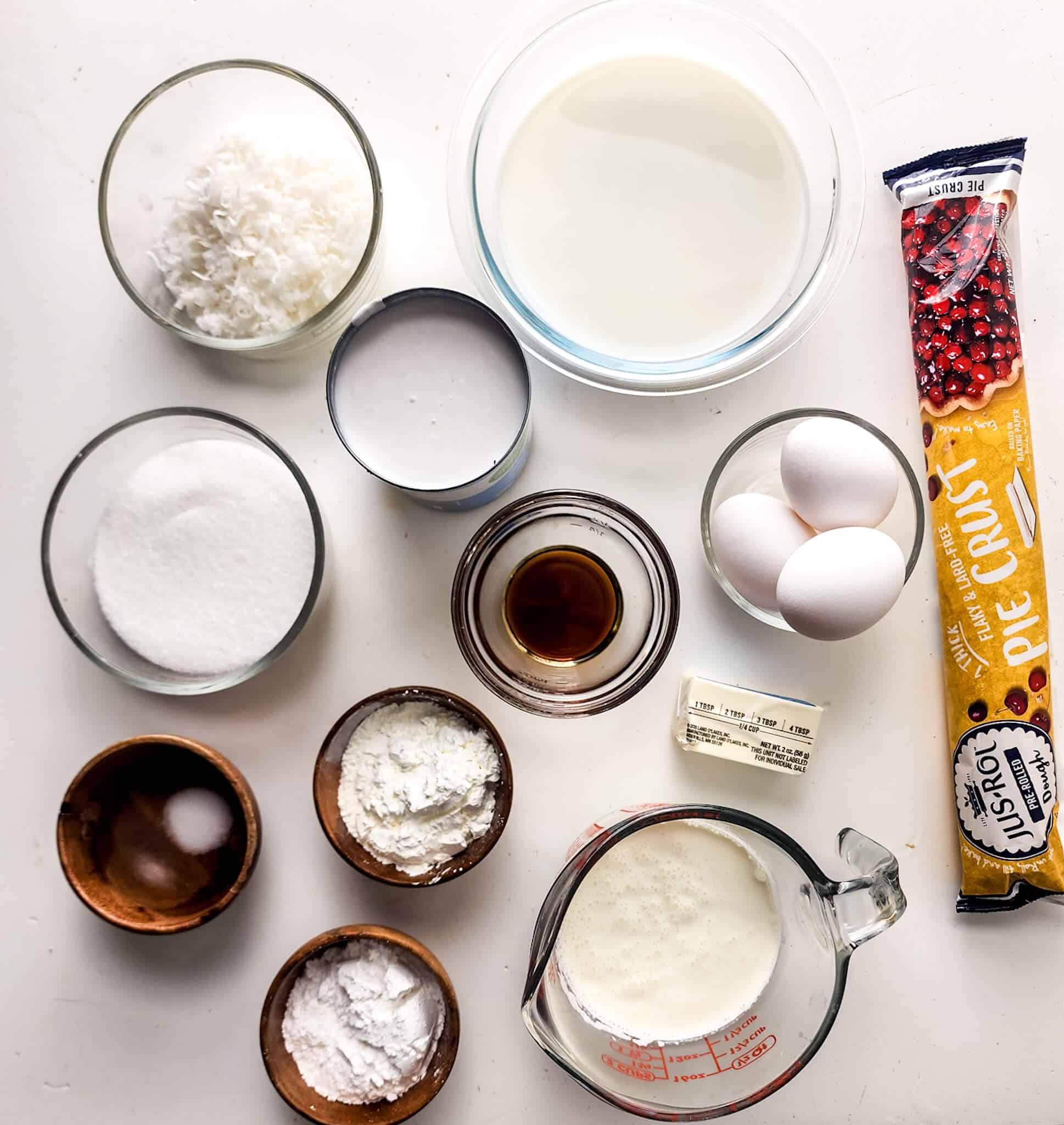 Ingredients for baking an old-fashioned coconut cream pie arranged on a white surface, including flour, milk, sugar, eggs, butter, and various other baking necessities.
