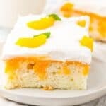 A square slice of Orange Creamsicle Cake topped with whipped cream, peach slices, and a mint leaf on a white plate.
