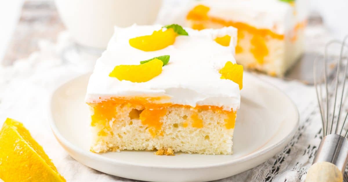A square slice of Orange Creamsicle Cake topped with whipped cream, peach slices, and a mint leaf on a white plate.