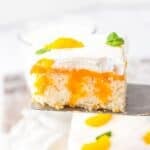 A slice of Orange Creamsicle Cake topped with whipped cream, orange filling, mandarin orange segments, and a mint leaf, held up on a spatula.