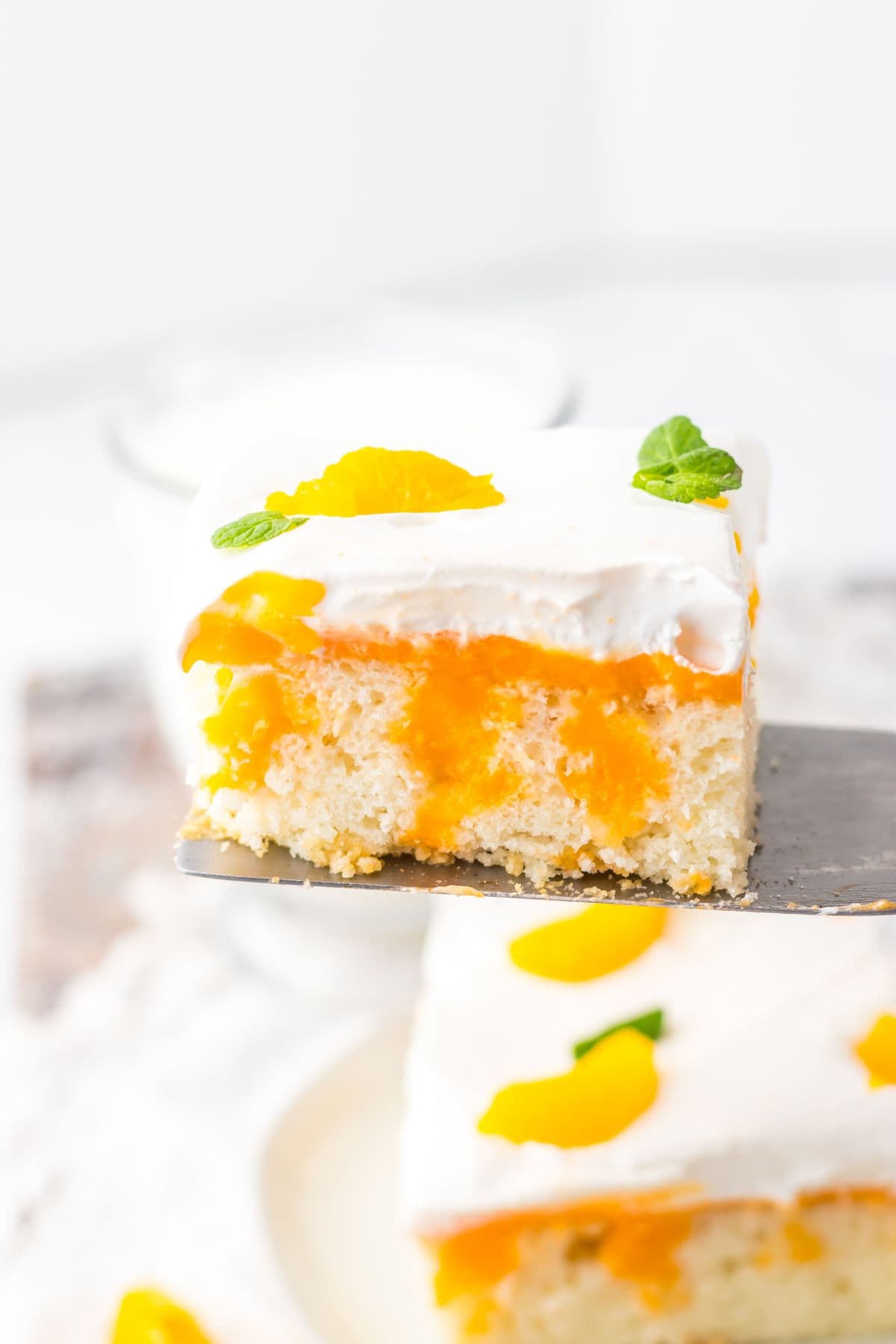 A slice of Orange Creamsicle Cake topped with whipped cream, orange filling, mandarin orange segments, and a mint leaf, held up on a spatula.