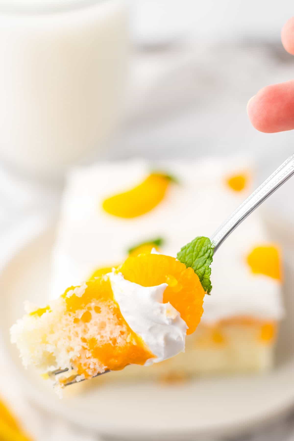 A fork holding a bite of Orange Creamsicle Cake topped with whipped cream, mandarin orange slices, and a mint leaf, with a glass of milk and cake slice in the background.