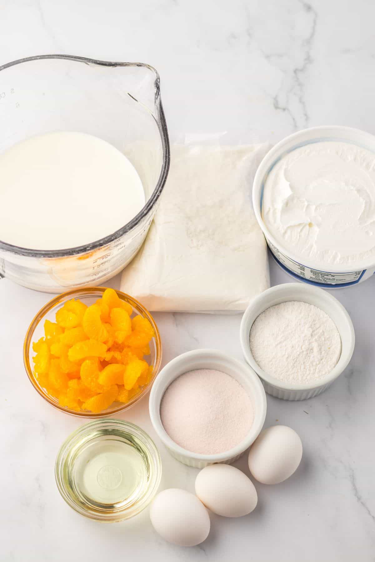 Ingredients  on a marble surface, including milk in a measuring cup, cream cheese, pastry dough, canned mandarin oranges, sugar, oil, flour, and three eggs.
