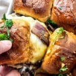 A hand lifts a cheesy, meat-filled slider topped with herbs from a dish of baked steak and cheese sliders.