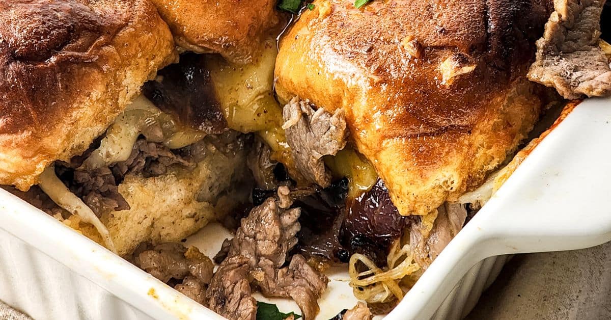 A close-up of a baked casserole with golden brown biscuits, melted cheese, sliced beef reminiscent of steak and cheese sliders, and caramelized onions in a white dish.
