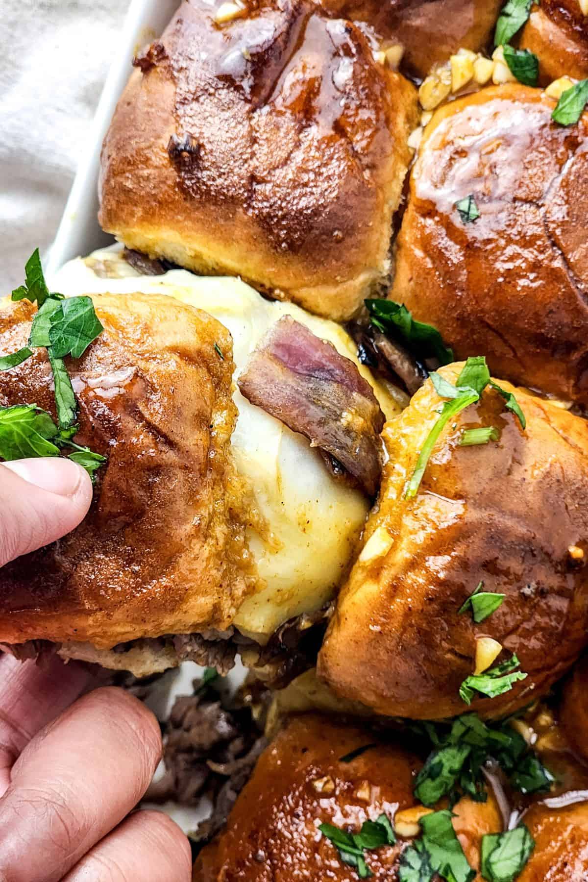 A hand lifts a cheesy golden bun, garnished with chopped parsley, from a tray.