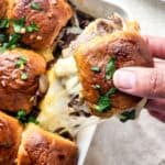 A hand lifts a cheesy steak and cheese slider topped with herbs from a baking dish filled with similar sliders.