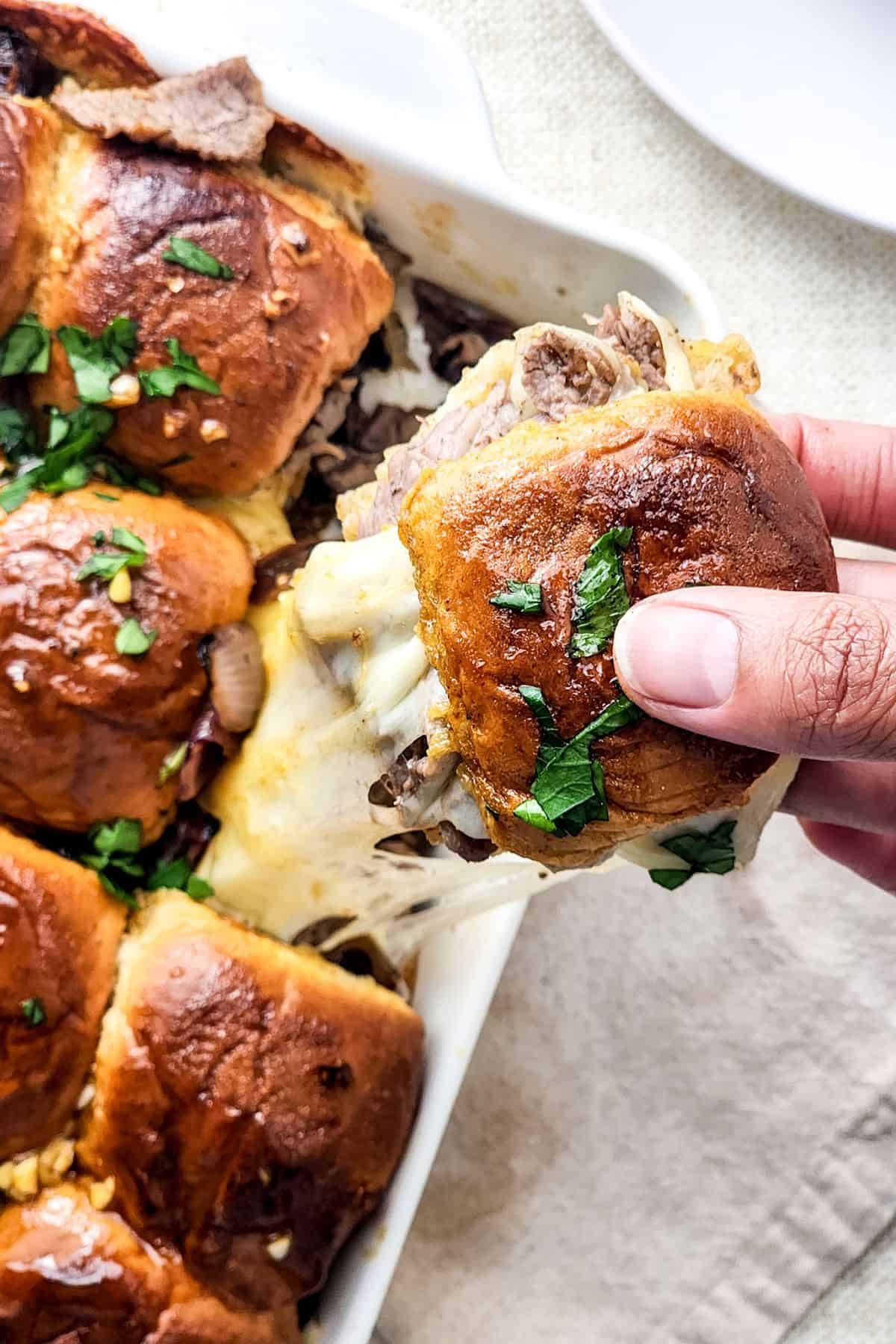 A hand lifts a cheesy steak and cheese slider topped with herbs from a baking dish filled with similar sliders.