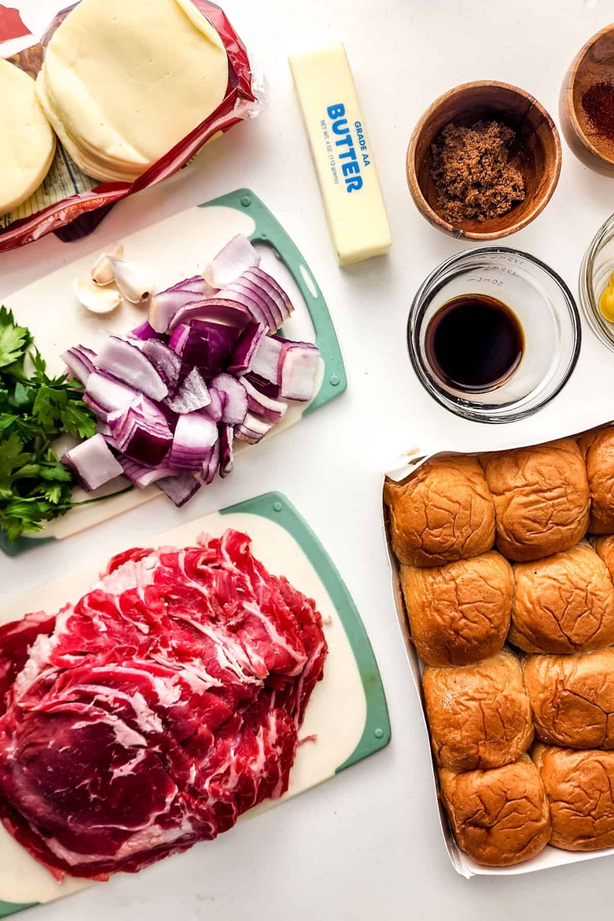Sliced provolone, butter, brown sugar, spices, Worcestershire sauce, parsley, chopped red onions, and sliced beef come together to create delicious steak and cheese sliders, with a tray of sandwich rolls arranged on a white surface.