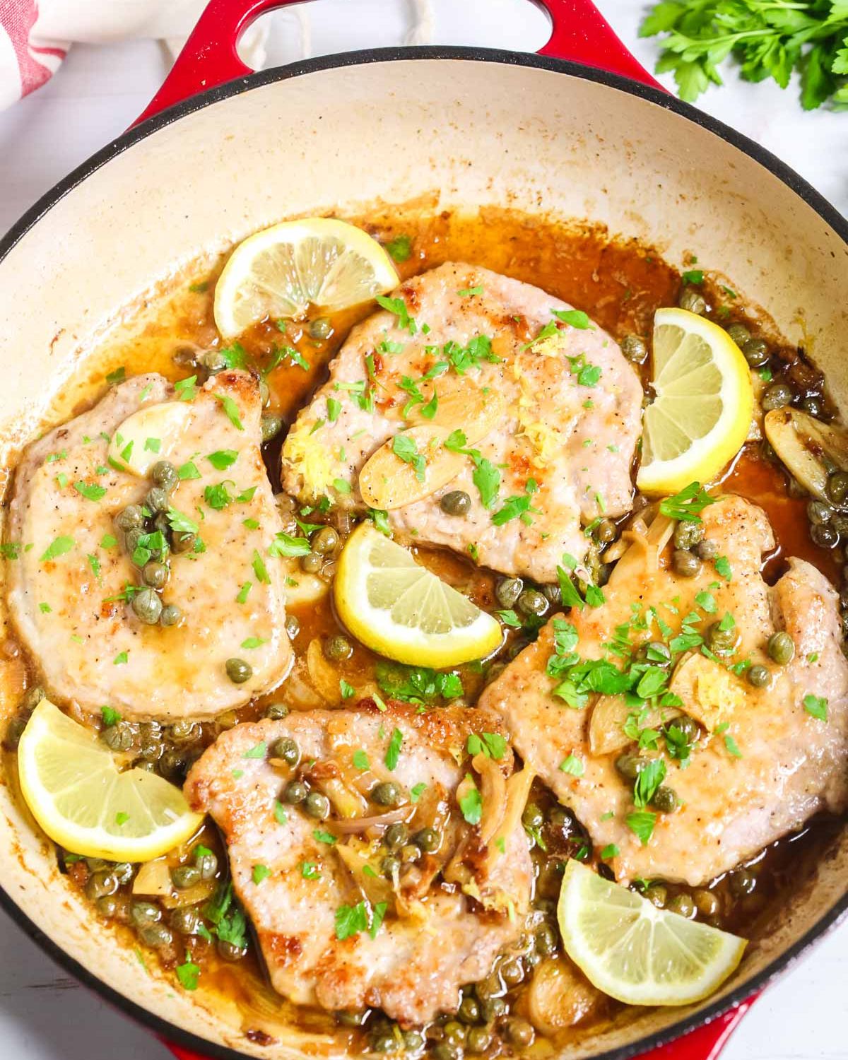 Pork piccata in a red skillet with lemons and parsley. 