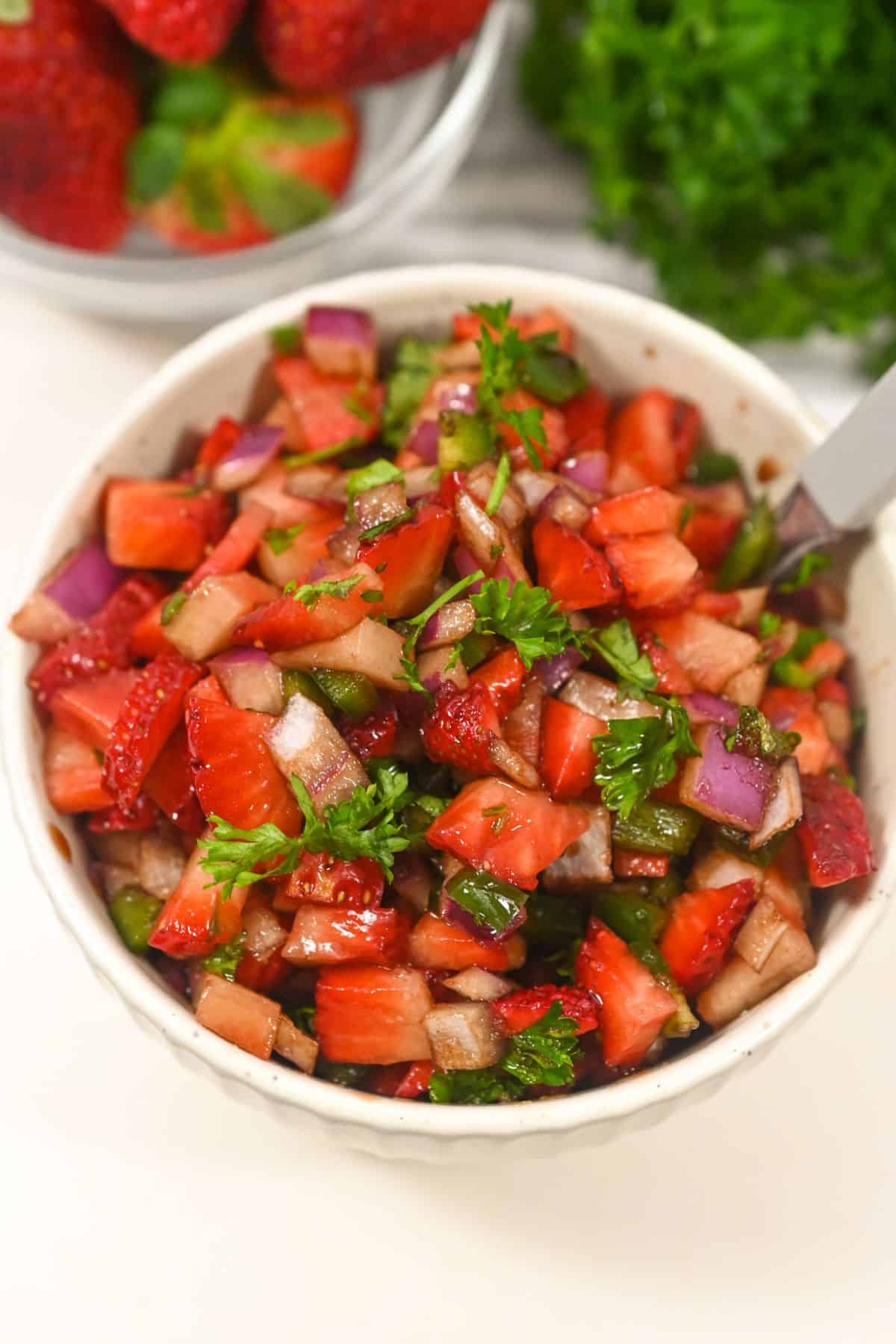 A vibrant bowl of strawberry salsa brimming with diced strawberries, red onion, green bell pepper, and fresh parsley sits alongside a fork; strawberries and herbs provide a colorful backdrop.