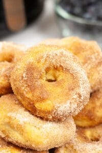 A close-up of a stack of cinnamon sugar-coated Air Fryer Biscuit Donuts, with a blurry bowl of blueberries in the background.