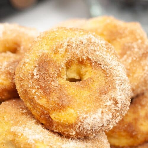 A close-up of a stack of cinnamon sugar-coated Air Fryer Biscuit Donuts, with a blurry bowl of blueberries in the background.