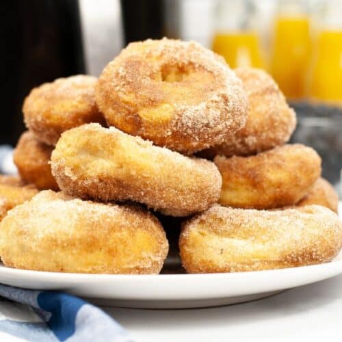 A plate of sugar-coated Air Fryer Biscuit Donuts stacked on a white dish, with bottles of orange juice and a blue checkered cloth in the background.