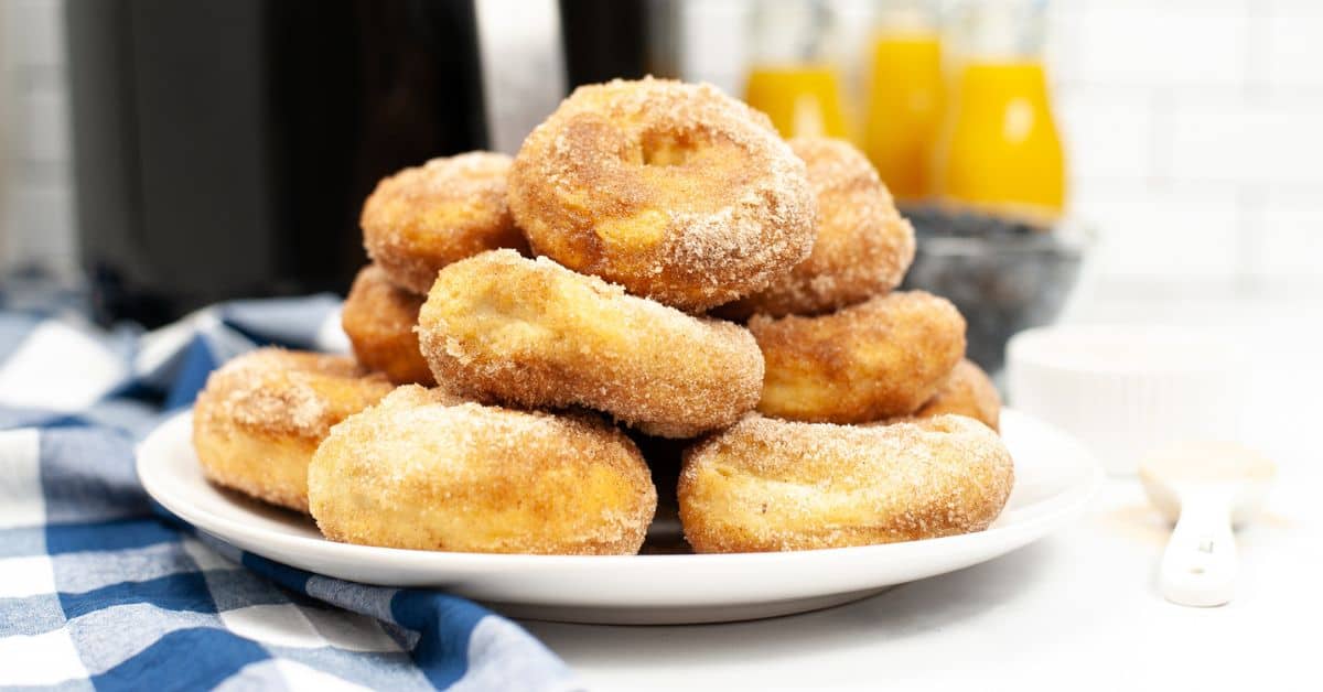 A plate of sugar-coated Air Fryer Biscuit Donuts stacked on a white dish, with bottles of orange juice and a blue checkered cloth in the background.