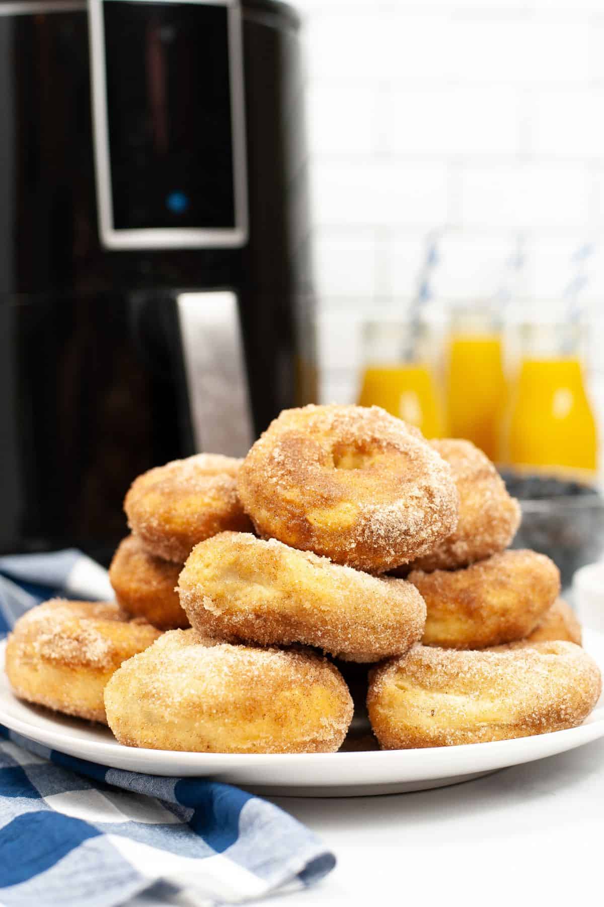 A plate of sugar-coated Air Fryer Biscuit Donuts is stacked in front of an air fryer, with bottles of orange juice and a blue checkered cloth nearby.