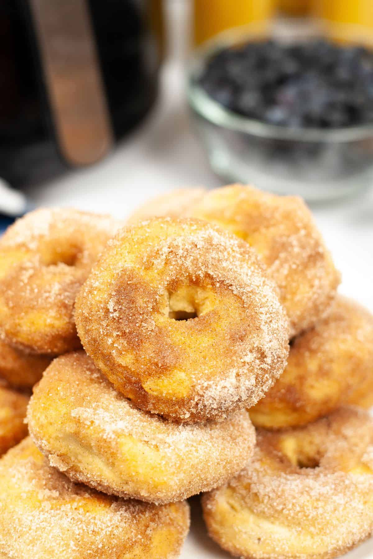 A stack of sugar-coated Air Fryer Biscuit Donuts is arranged on a white surface, with a bowl of blueberries and a coffee pot blurred in the background.