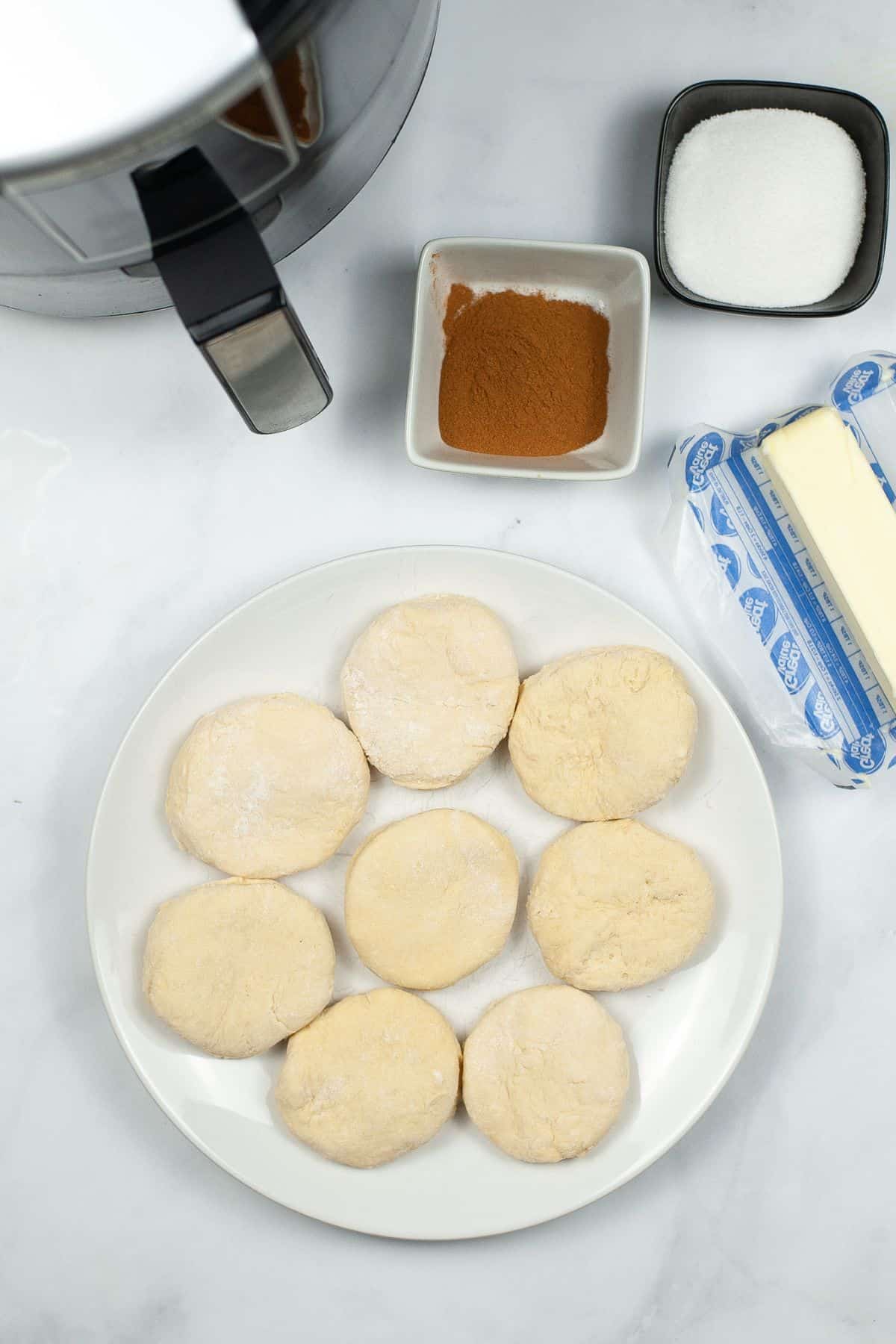 A plate of eight unbaked biscuit dough rounds, butter, cinnamon, and sugar is ready on a white countertop next to an air fryer—perfect for making delicious Air Fryer Biscuit Donuts.