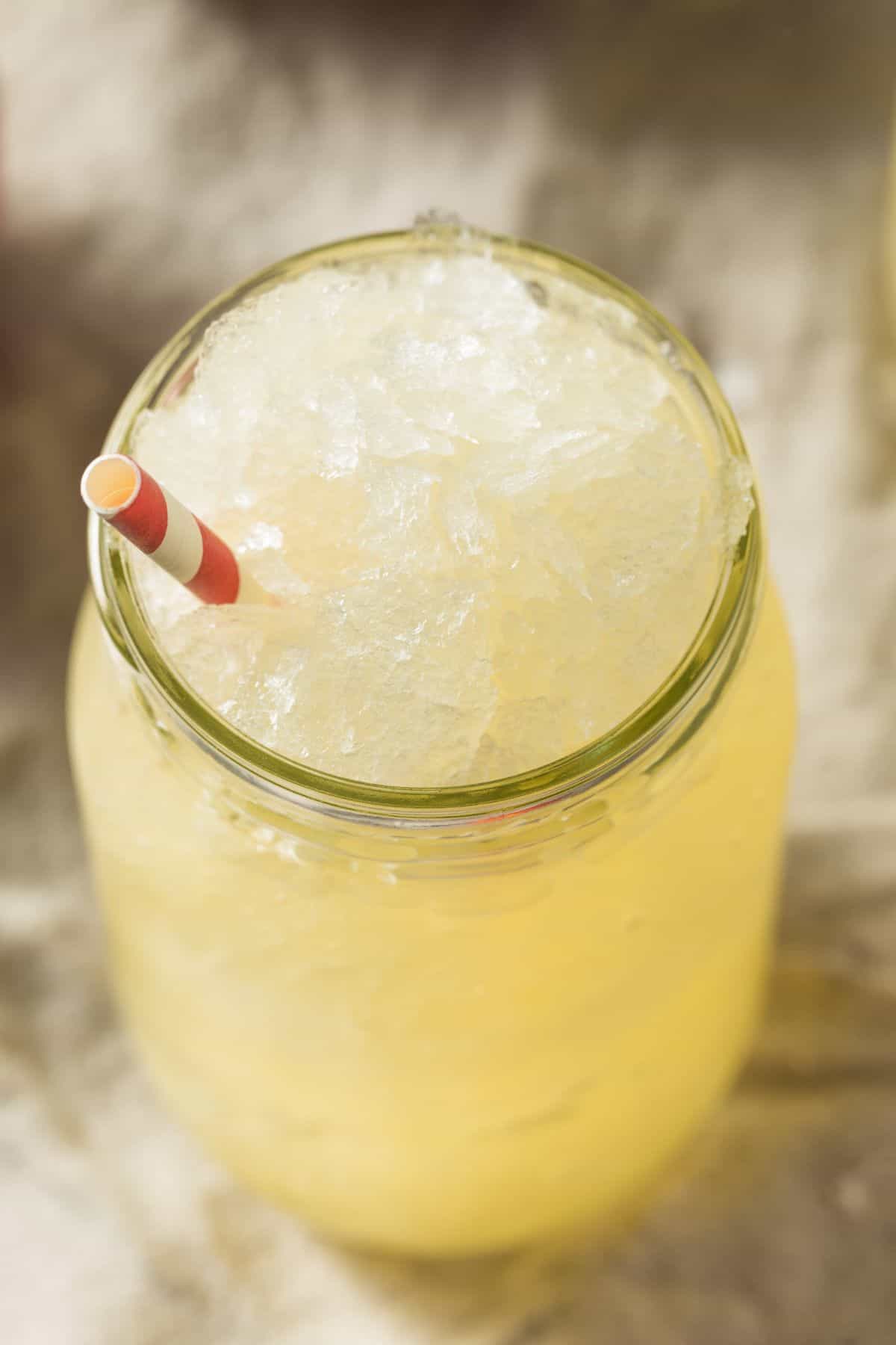 A mason jar filled with a refreshing drink its yellow iced drink topped with crushed ice and a red and white striped straw, sits invitingly on a neutral surface.