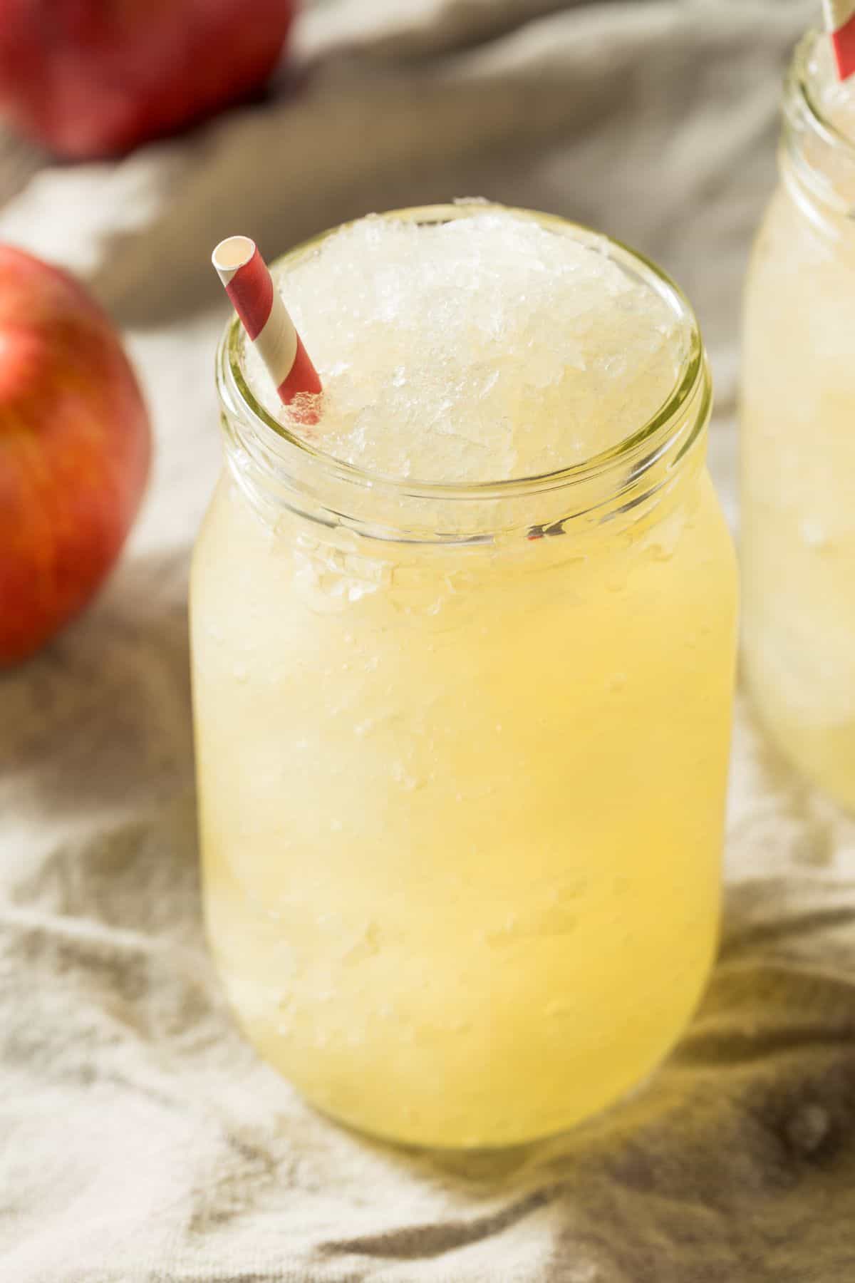 A mason jar filled with refreshing Apple Cider Slush and a red and white striped straw sits on a cloth surface, with an apple in the background.