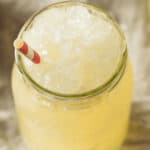 Glass jar filled with crushed ice and a yellow Apple Cider Slush, topped with a red and white striped paper straw.