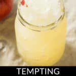 A mason jar filled with refreshing apple cider slush and a red-striped straw on a light surface, an apple in the background. Text at the bottom reads "Tempting Apple Cider Slush.
