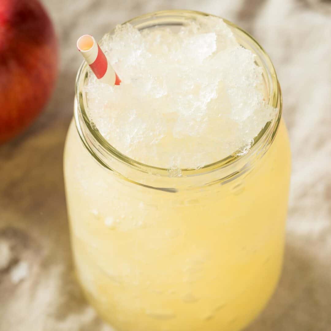 A glass jar filled with a yellow Apple Cider Slush and ice, topped with a red and white striped straw. An apple is partially visible in the background.
