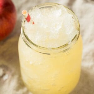 A glass jar filled with a yellow Apple Cider Slush and ice, topped with a red and white striped straw. An apple is partially visible in the background.