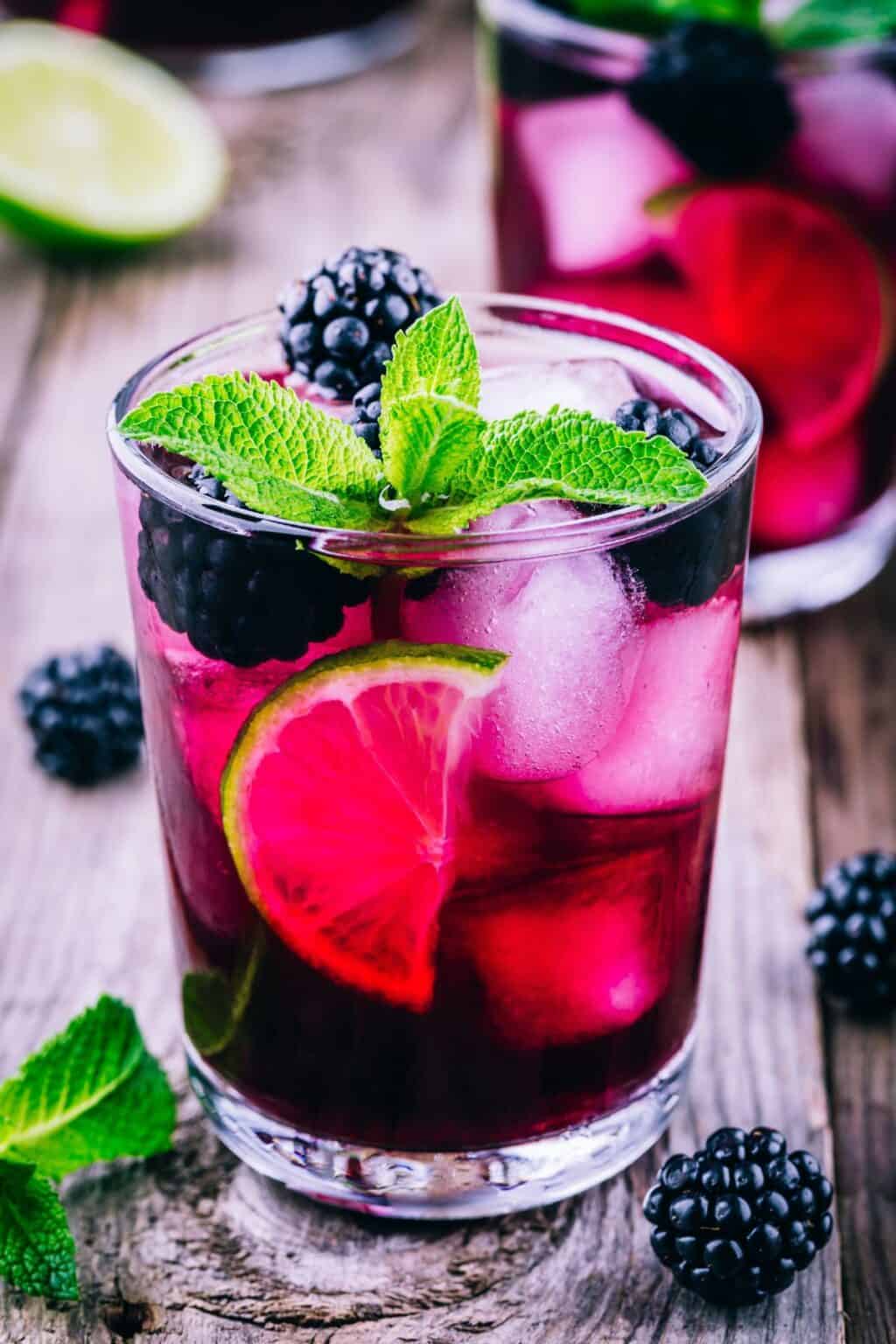 A refreshing glass of Blackberry Bourbon Smash, topped with blackberries, lime slices, and a sprig of mint, sits invitingly on a wooden surface.