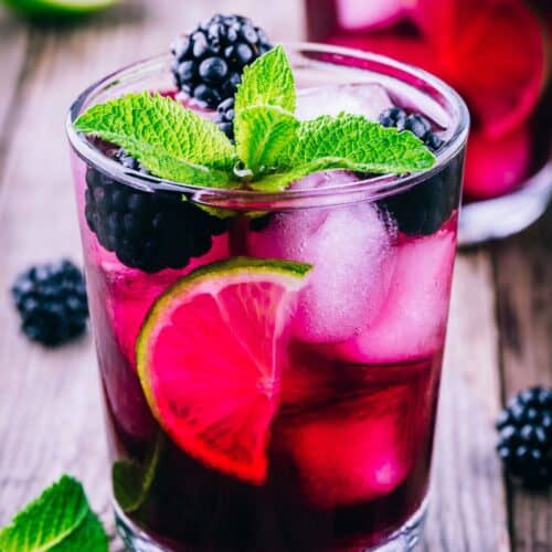 A refreshing glass of Blackberry Bourbon Smash, topped with blackberries, lime slices, and a sprig of mint, sits invitingly on a wooden surface.