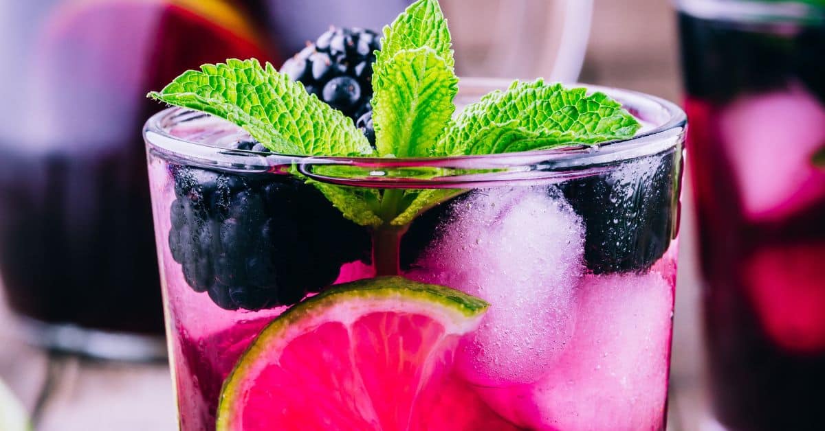 A glass of purple Blackberry Bourbon Smash with ice, blackberries, a mint sprig, and a slice of lime.