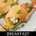 Breakfast croissant roll-ups filled with scrambled eggs, tomatoes, spinach, green onions, and cheese, topped with sesame seeds and green chili slices. Presented on a baking sheet with the text reading "Delicious Breakfast Croissant Roll-Ups.