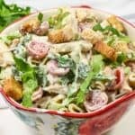 A bowl with penne, sliced grape tomatoes, lettuce, croutons, and a white dressing, served in a floral-patterned bowl.