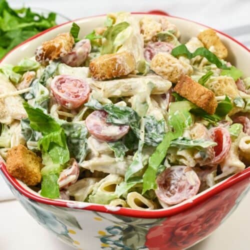 A bowl with penne, sliced grape tomatoes, lettuce, croutons, and a white dressing, served in a floral-patterned bowl.