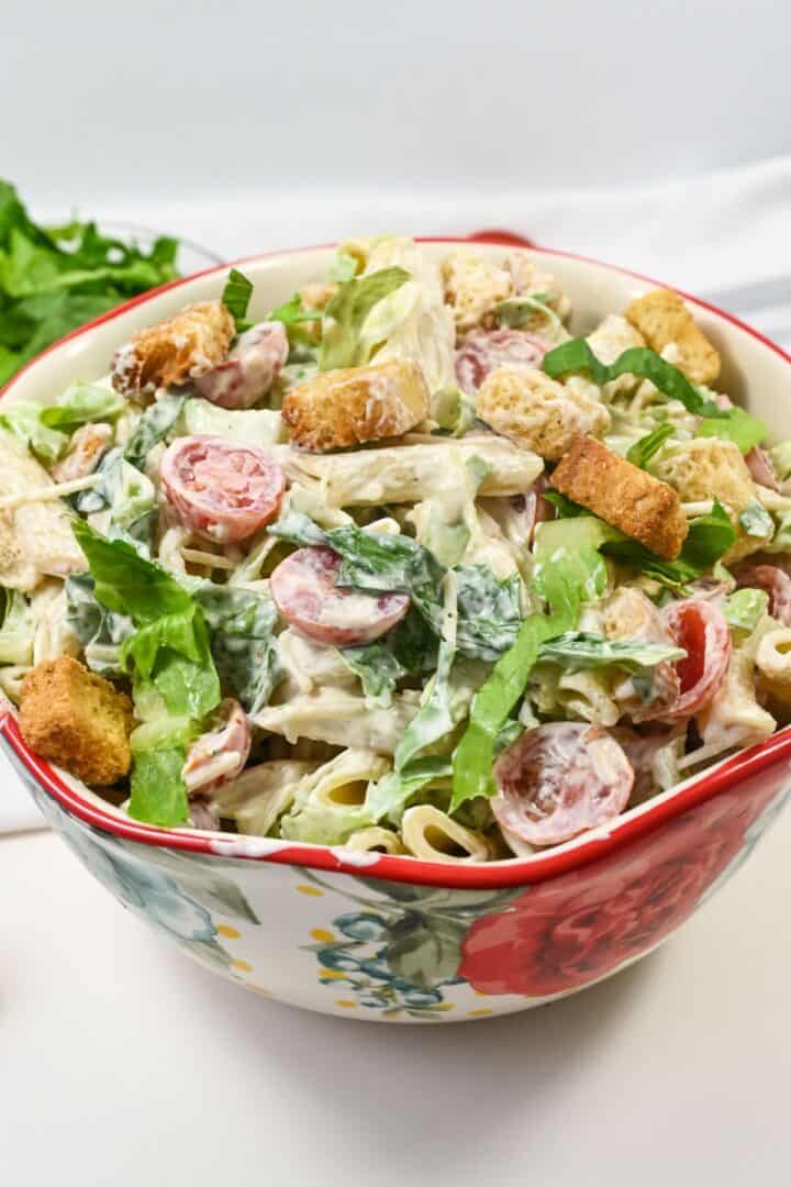 A bowl with penne, sliced grape tomatoes, lettuce, croutons, and a white dressing, served in a floral-patterned bowl.