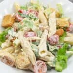 A plate of Caesar Pasta Salad with penne, lettuce, cherry tomatoes, croutons, and creamy dressing on a white plate.