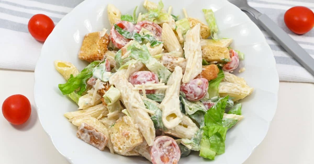 A plate of Caesar Pasta Salad with penne, lettuce, cherry tomatoes, croutons, and creamy dressing on a white plate.