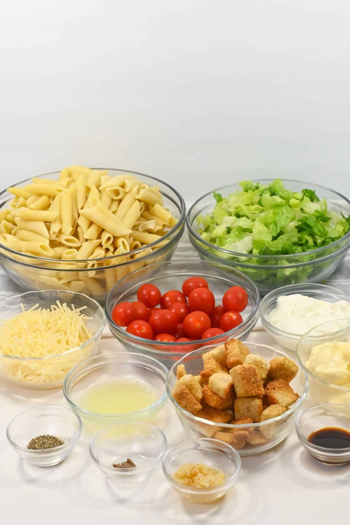 Various salad ingredients in glass bowls, including penne pasta, chopped lettuce, cherry tomatoes, shredded cheese, croutons, mayonnaise, seasoning, lemon juice, and minced garlic.